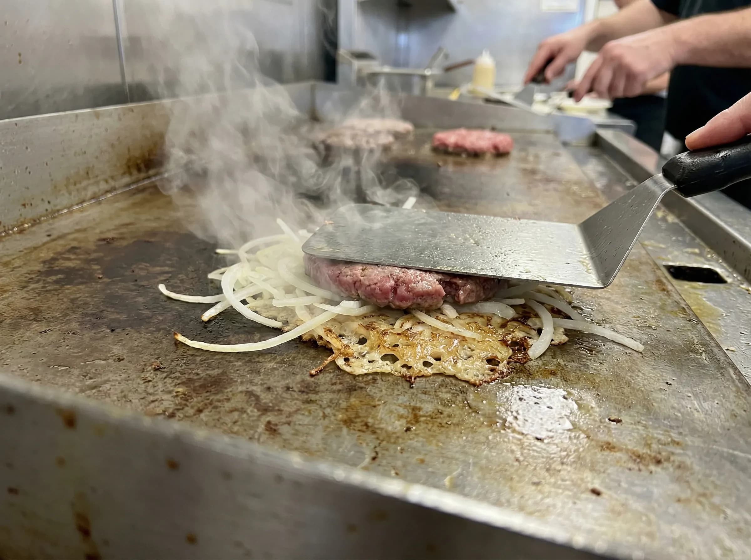 Oklahoma-style onion burger being smashed into thinly sliced onions on a flat-top griddle
